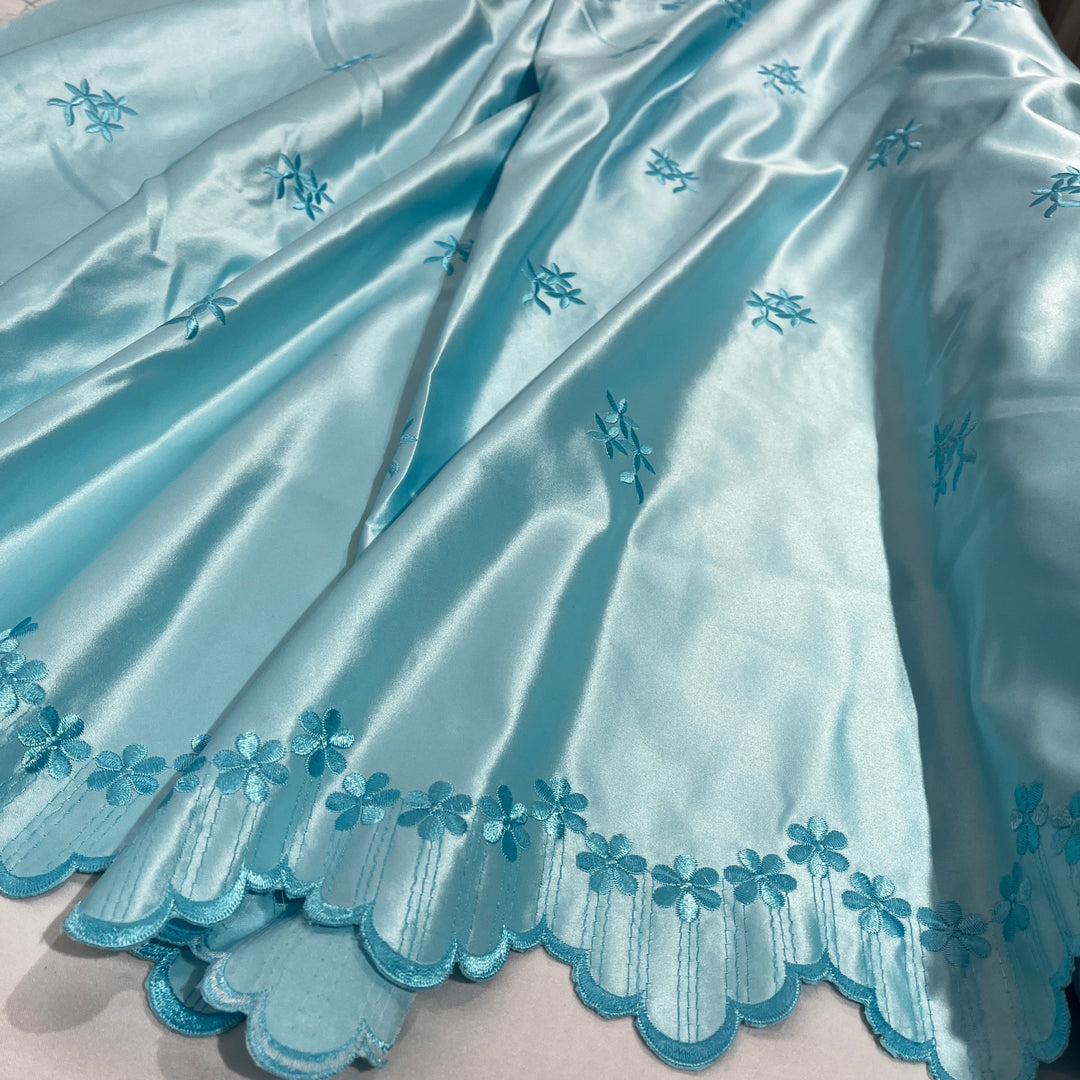 Aua blue satin fabric with embroidered floral motifs and a scalloped embroidered border, shown in a close-up draped view.