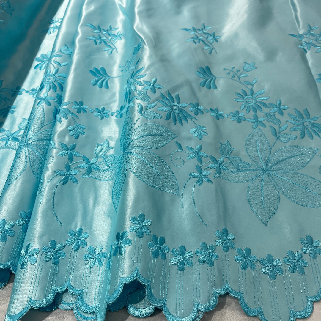 A close-up of a light aqua blue satin fabric with detailed floral embroidery and a scalloped edge.