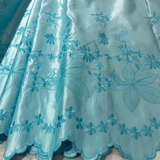 A close-up of a light aqua blue satin fabric with detailed floral embroidery and a scalloped edge.