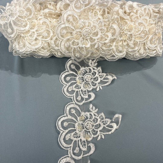 Ivory floral lace trim embellished with embroidered flowers, pearls, and sequins, featuring a scalloped edge and intricate floral pattern., draped over a gray fabric background. LaceUSA #KZ-50