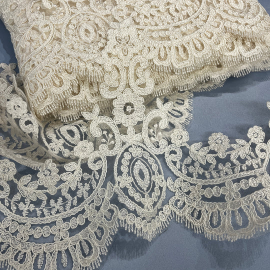 Close-up of ivory floral lace trim with ornate embroidery and scalloped edging, laid on matching lace against a slate blue background, Lace USA 96794W.