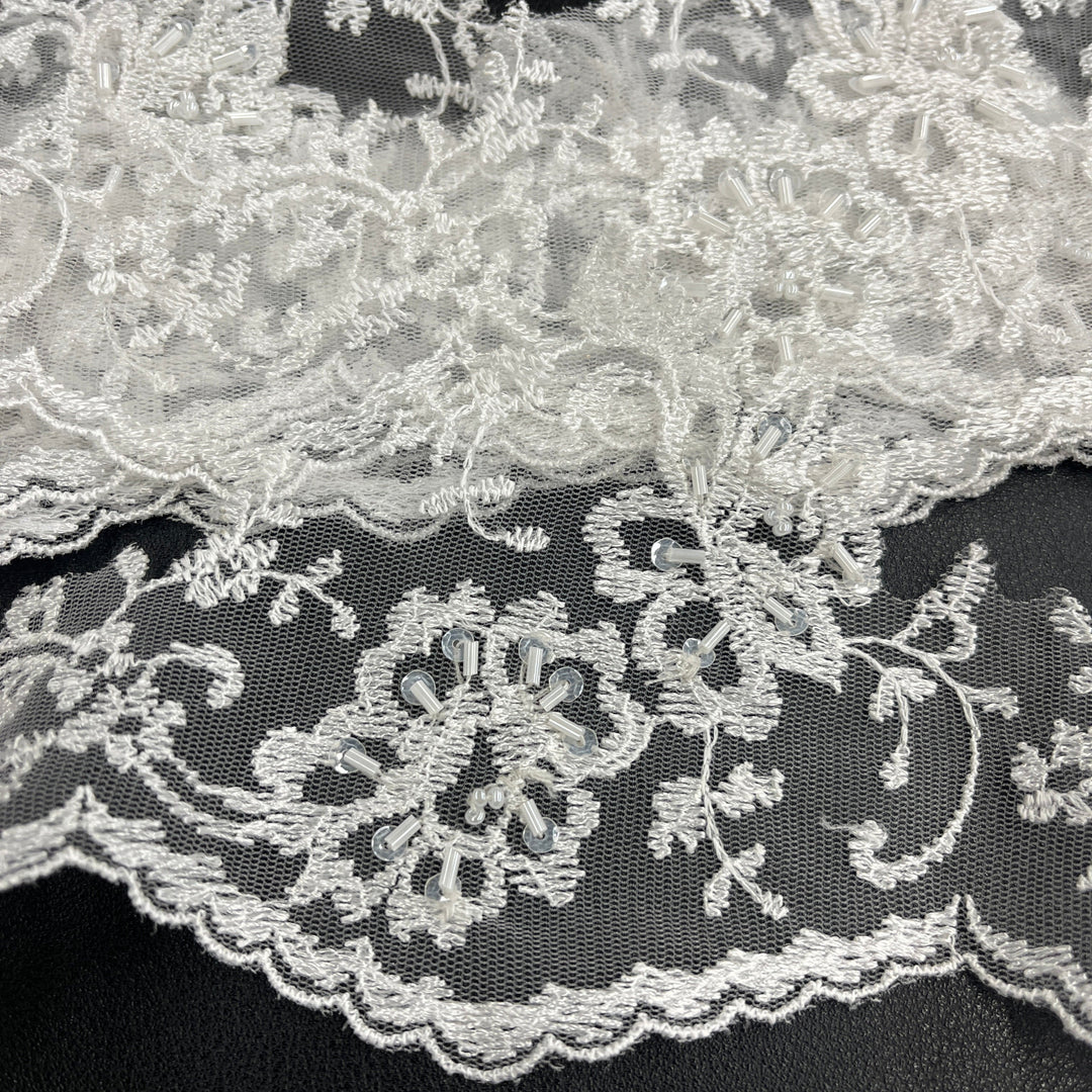 Close-up of white beaded floral lace trim with a scalloped edge and embroidered floral motifs on fine net mesh, layered over itself against a black background. LaceUSA #21356WBP