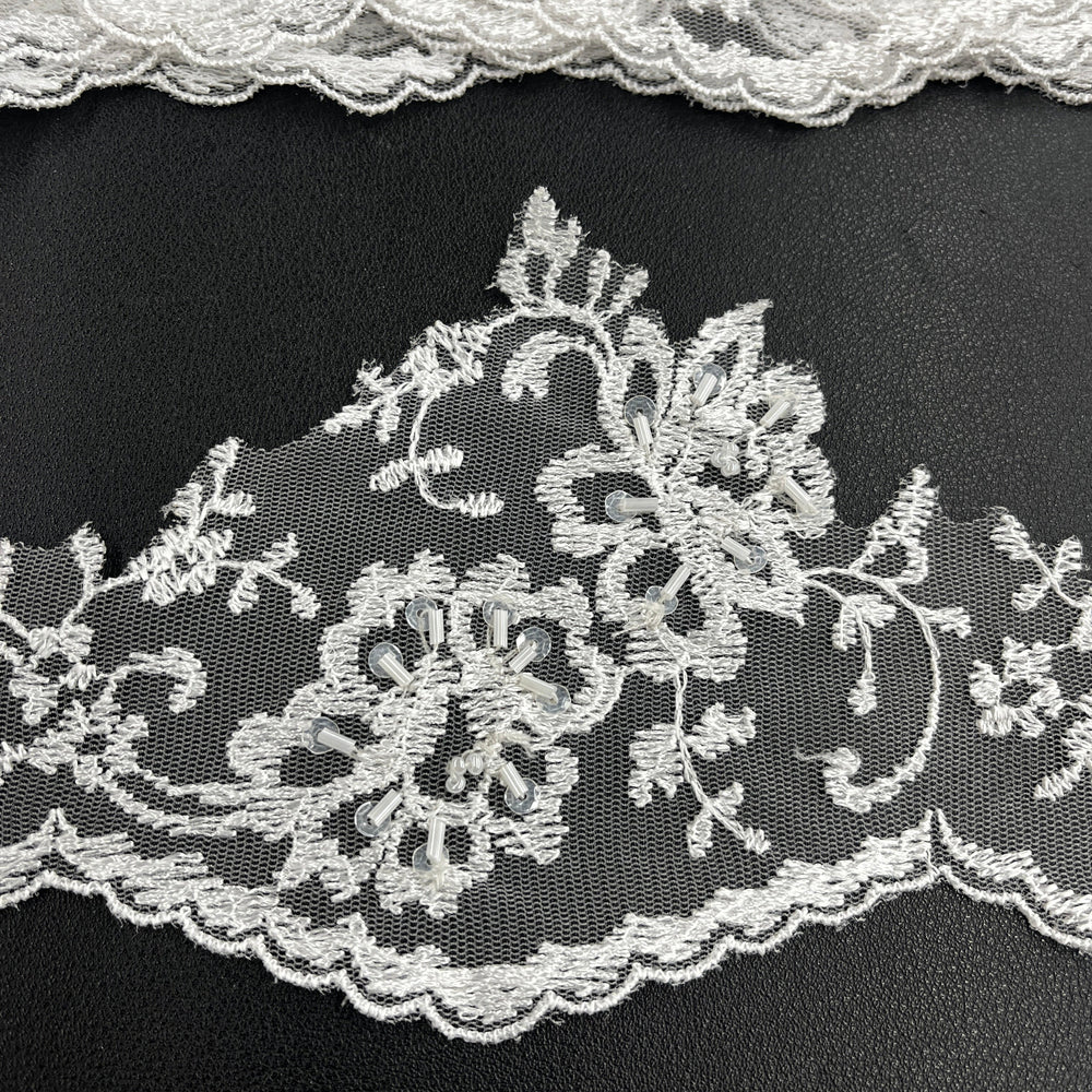 Closeup of a white floral lace trim with scalloped edge, embellished with beads and sequins, displayed on a black background. LaceUSA #21356WBP