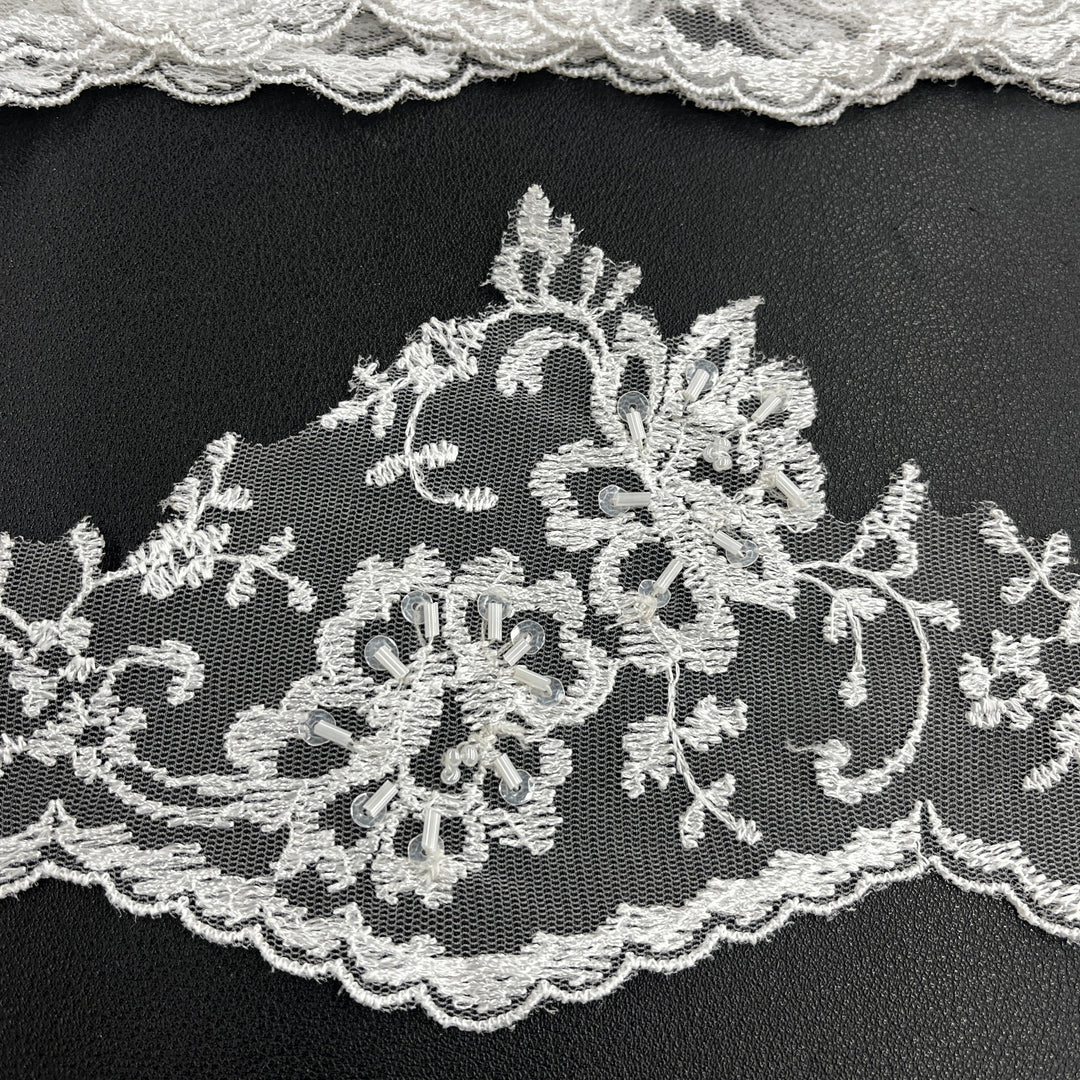 Closeup of a white floral lace trim with scalloped edge, embellished with beads and sequins, displayed on a black background. LaceUSA #21356WBP