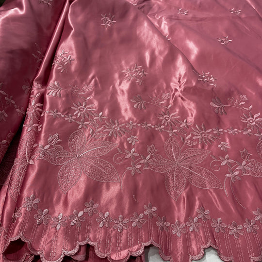 A close-up of a dusty rose satin fabric with intricate floral and leaf embroidery and a scalloped border.