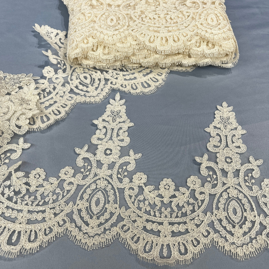 Ivory corded floral lace trim with ornate embroidery and scalloped edging, shown laid out on a slate blue background with a folded piece of the same lace at the top. Lace USA 96794W