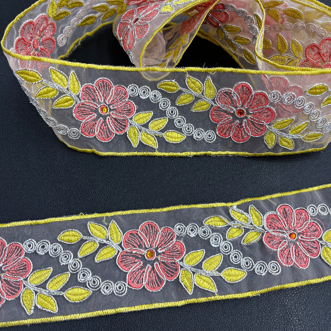 Close-up of two pieces of the same decorative embroidered lace trim, featuring pink flowers with rhinestone centers, yellow leaves, and silver scroll details, corded with metallic silver thread on sheer organza fabric over a dark surface. Lace USA #96307NRS. 