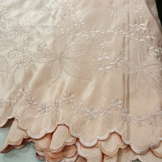 A close-up of a peach satin fabric with intricate floral embroidery and a scalloped embroidered border.