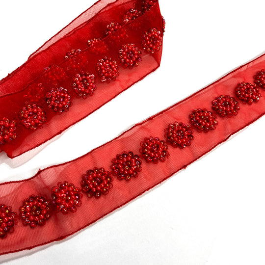Close-up of red sheer net mesh trim ribbon with beaded and sequined floral patterns, shown coiled and laid flat diagonally on a white background. Lace USA #21766WNBP