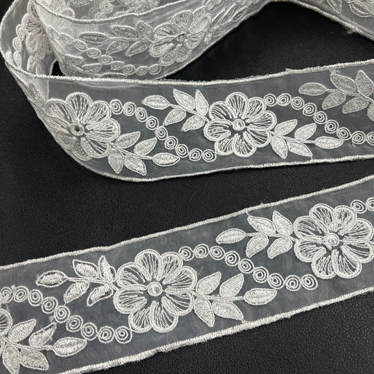 Close-up of two pieces of the same  silver decorative embroidered lace trim, featuring flowers with rhinestone centers, leaves, and scroll details, corded with metallic silver thread on sheer organza fabric over a dark surface. Lace USA #96307NRS. 