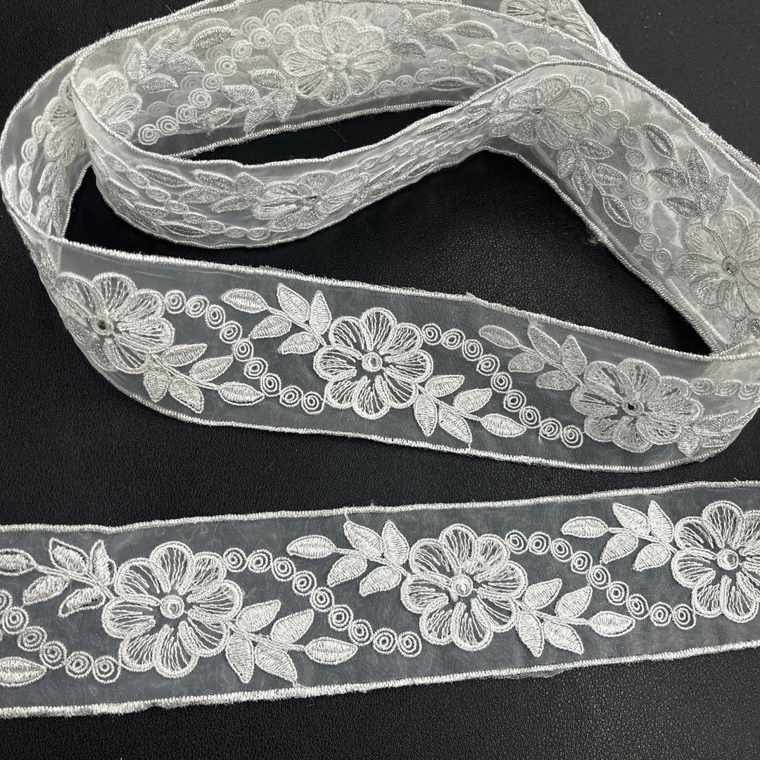 Two pieces of the same silver decorative embroidered lace trim, featuring flowers with rhinestone centers, leaves, and scroll details, corded with metallic silver thread on sheer organza fabric over a dark surface. Lace USA #96307NRS. 