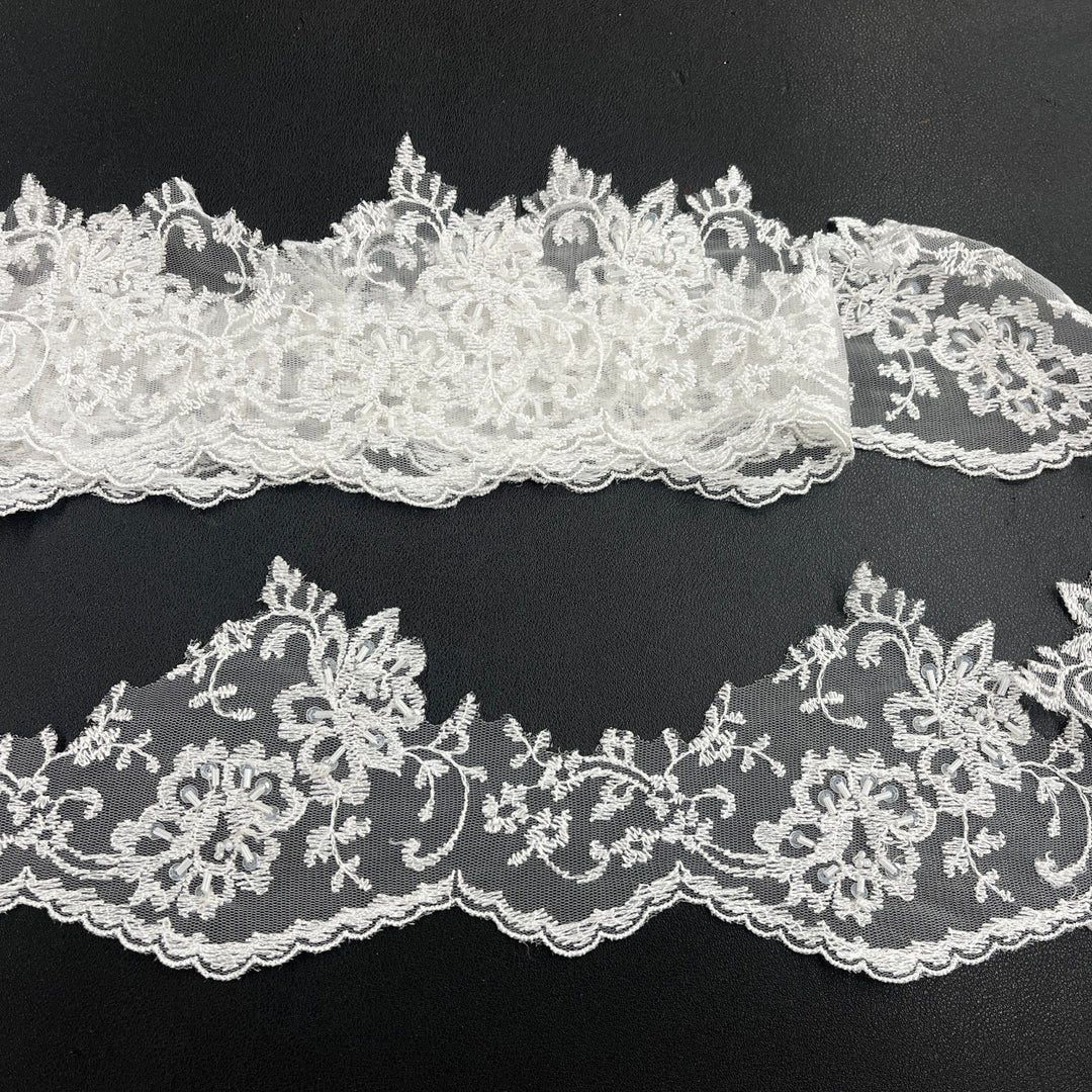 White beaded floral lace trim with scalloped edges, displayed in two horizontal strips featuring one flat unrolled length and one gathered bundle on a black background. Lace_USA #21356WBP
