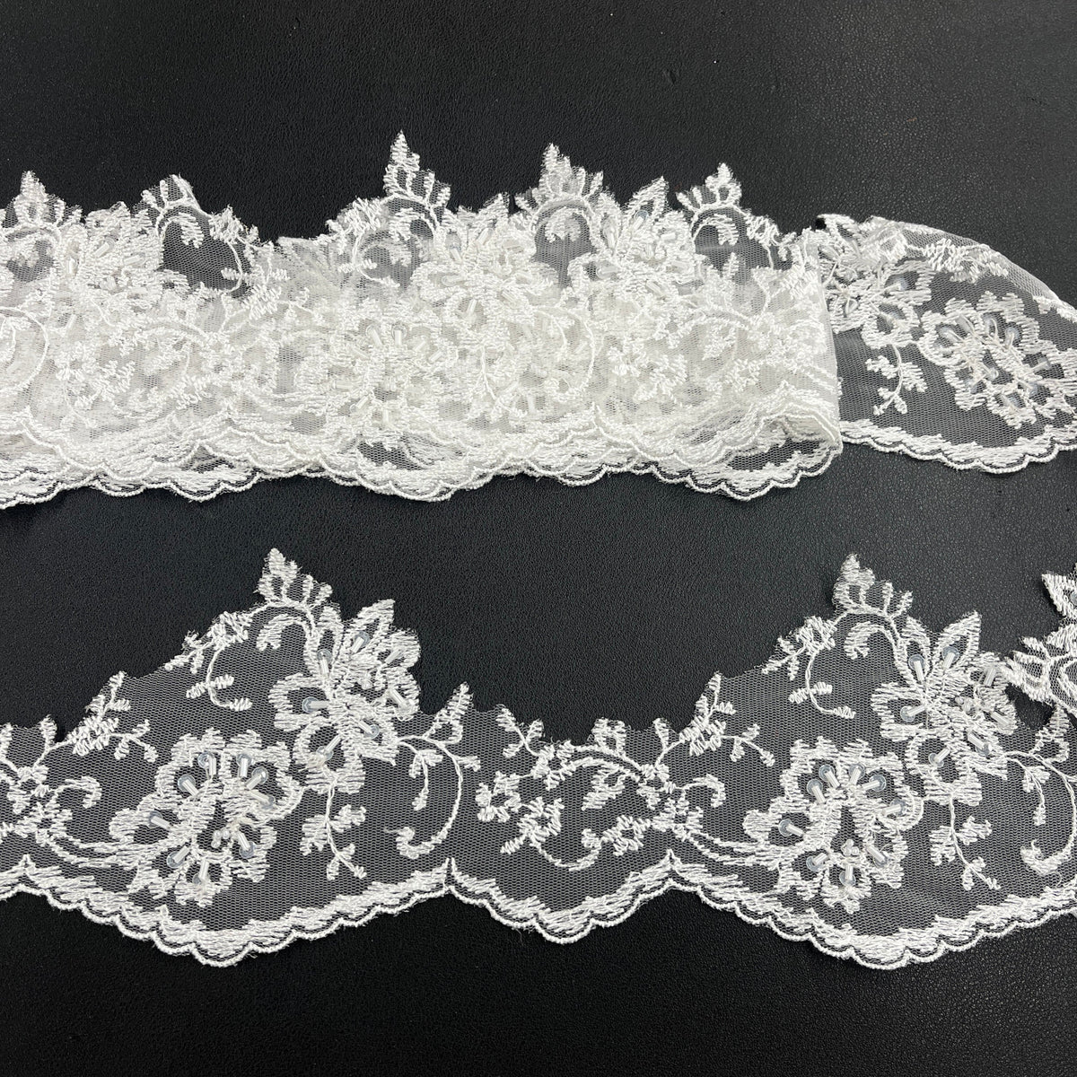 White beaded floral lace trim with scalloped edges, displayed in two horizontal strips featuring one flat unrolled length and one gathered bundle on a black background. Lace_USA #21356WBP
