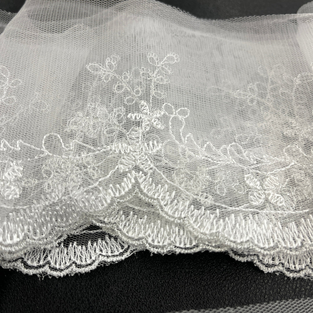 White floral lace trim with embroidered details and a scalloped tulle edge, shown bunddled against a dark background. Lace USA 21734W