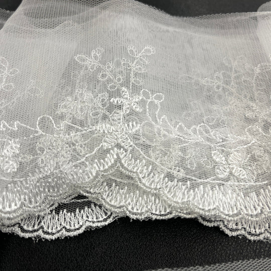 White floral lace trim with embroidered details and a scalloped tulle edge, shown bunddled against a dark background. Lace USA 21734W