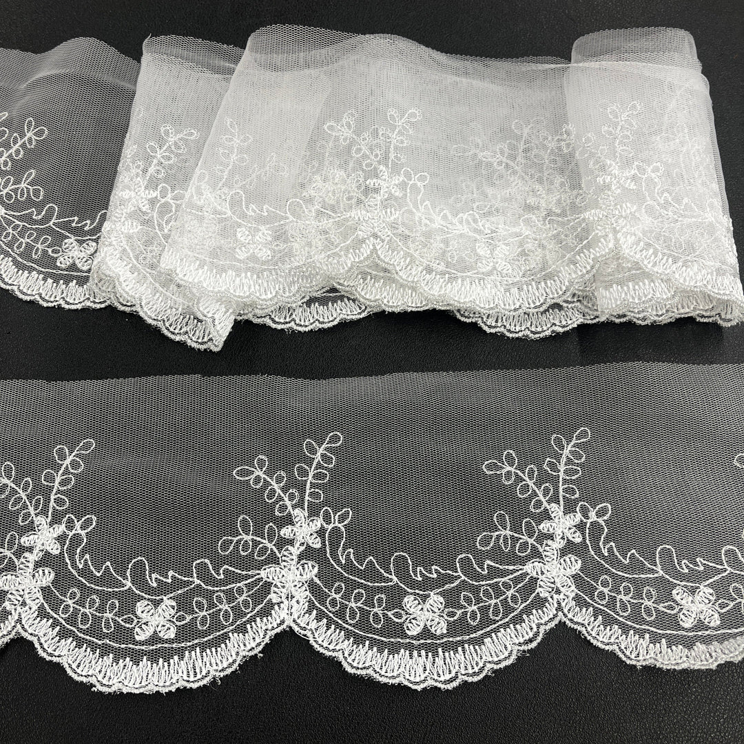 White floral lace trim with scalloped edges, embroidered on 100% polyester net mesh,, featuring one flat unrolled length and one gathered bundle on a black background. Lace USA 21734W