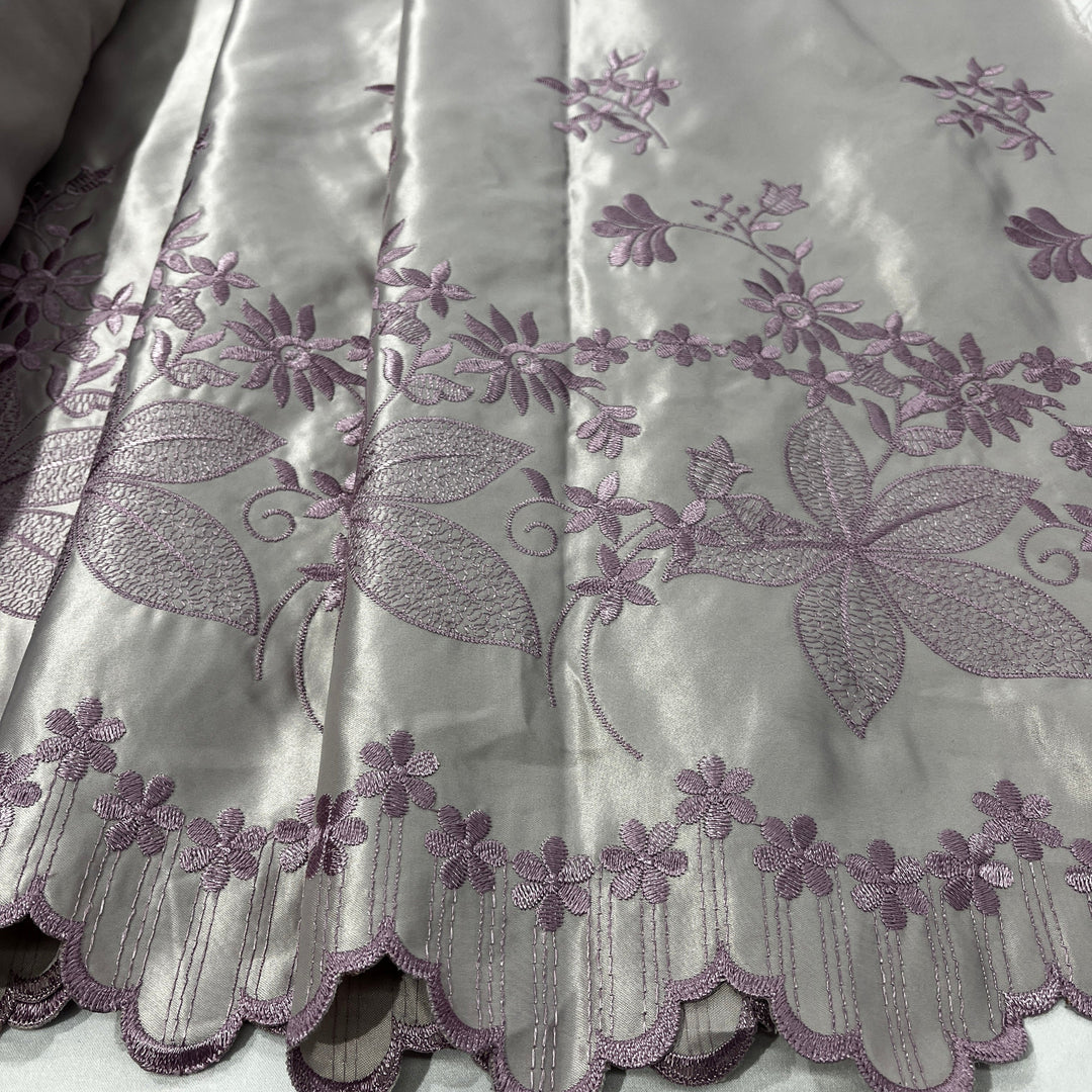 A close-up of a yam color fabric with lavender-purple floral embroidery and a scalloped embroidered border.