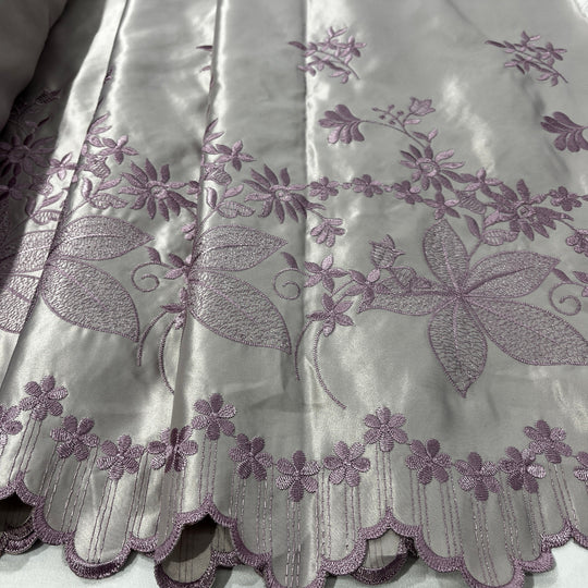A close-up of a yam color fabric with lavender-purple floral embroidery and a scalloped embroidered border.
