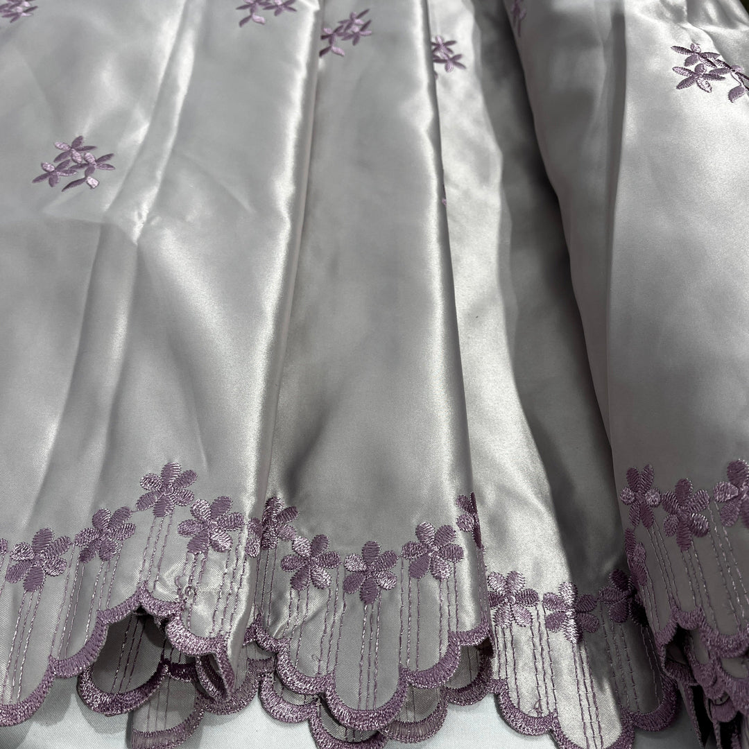 A close-up of a yam satin fabric with a scalloped hem and lavender-purple floral embroidery.