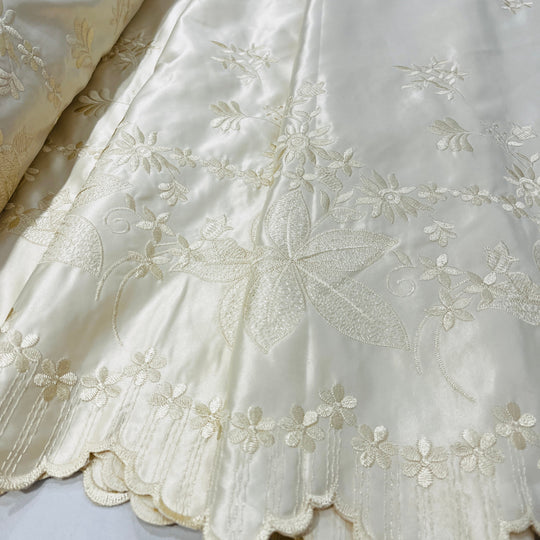Close-up of an champagne satin fabric with delicate floral embroidery and a scalloped embroidered border.