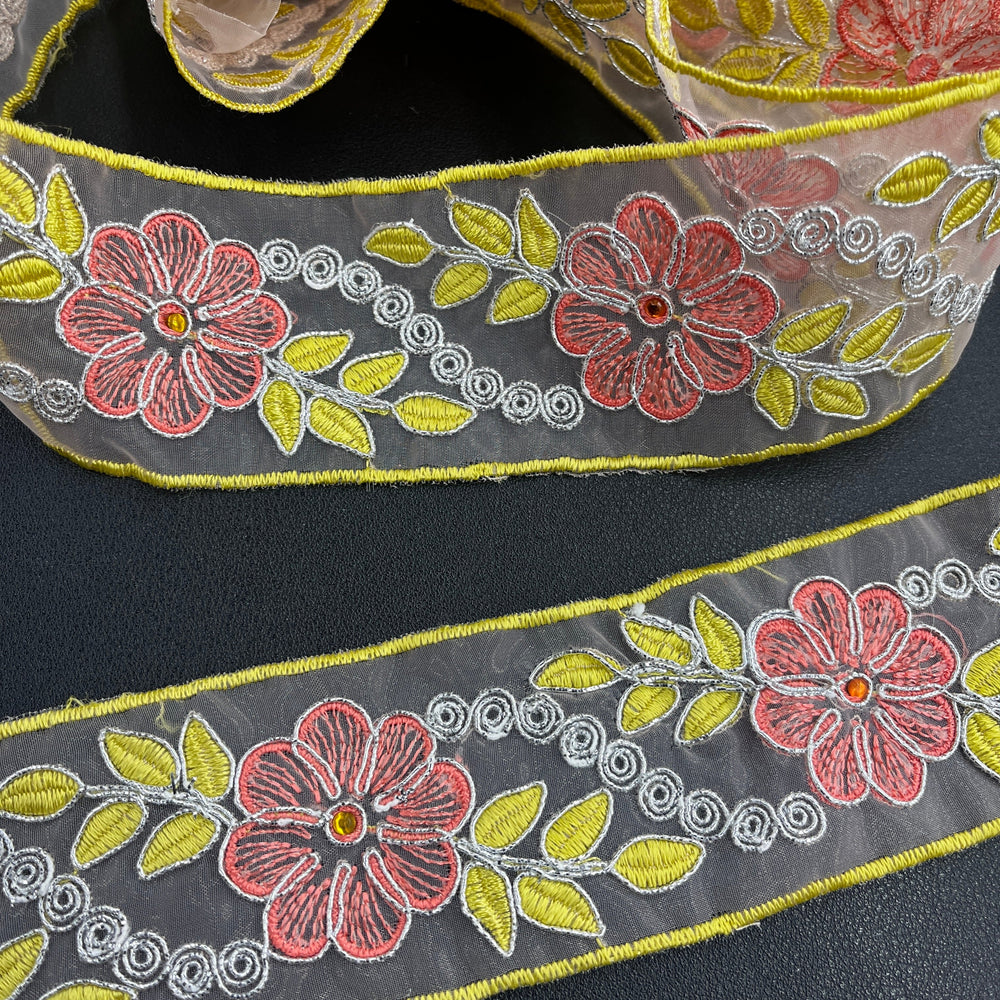 Close-up of two pieces of the same decorative embroidered lace trim, featuring pink flowers with rhinestone centers, yellow leaves, and silver scroll details, corded with metallic silver thread on sheer organza fabric over a dark surface. Lace USA #96307NRS. 