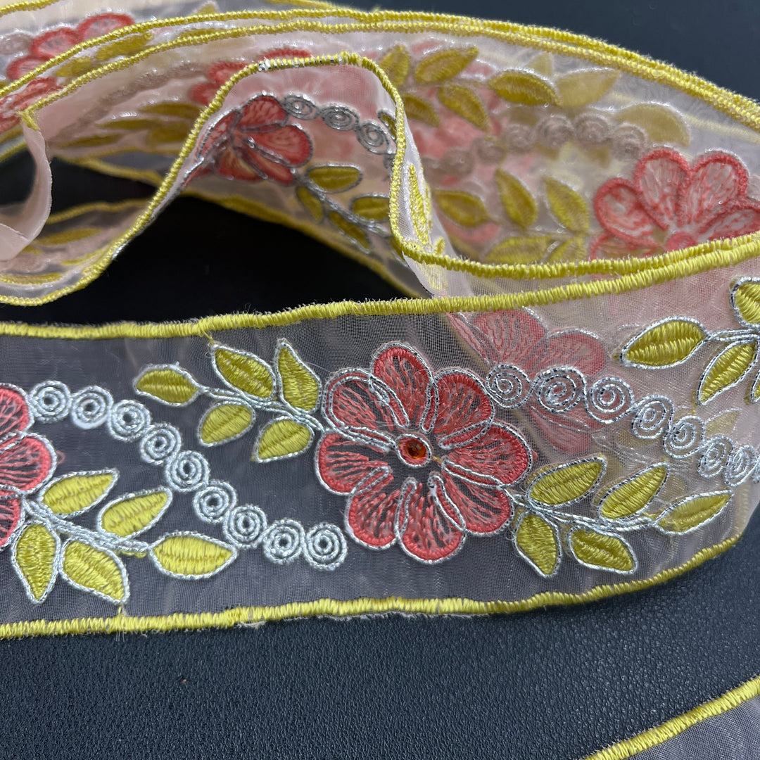 Close-up of the a decorative embroidered lace trim, featuring pink flowers with rhinestone centers, yellow leaves, and silver scroll details, corded with metallic silver thread on sheer organza fabric over a dark surface. Lace USA #96307NRS. 